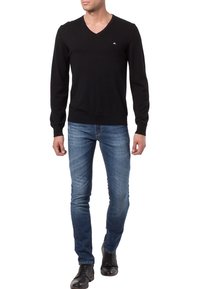 Maglione nero a maglia con scollo a V e maniche lunghe, indossato con jeans azzurri chiari e stivali in pelle nera. Presenta un piccolo logo sul petto.