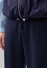 Giacca leggera blu navy con orlo arricciato, dotata di una cerniera argento e cordini, abbinata a pantaloni blu navy coordinati. Texture morbida.