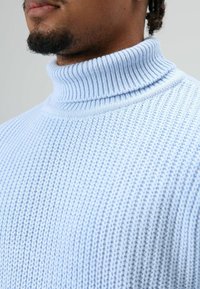 Pull à col roulé bleu clair en maille avec un motif texturé et un col côtelé, présentant un gros plan du tissu et des détails du design.