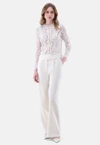 Top a maniche lunghe in pizzo con motivi floreali trasparenti, abbinato a pantaloni bianchi fluente a vita alta. Il modello è in piedi con le mani in tasca.