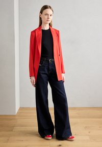Blazer rosso, top nero, jeans in denim scuri a gamba larga e scarpe con plateau rosse. Tessuto liscio, spalle strutturate e linee pulite.
