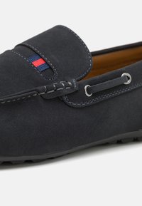 Námořnické modré semišové mokasíny se šitým designem, s malým červeným a modrým pruhem a kovovými očky na tkaničky. Texturovaná gumová podrážka.