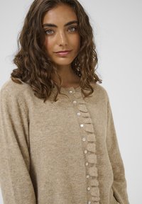 Mlada ženska z valovitimi rjavimi lasmi nosi bež pleteno jopico z bisernimi gumbi in volančki spredaj, proti enotni ozadju.