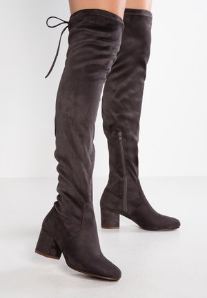 Ciemnobrązowe zamszowe kozaki do uda na obcasie typu słupek, z bocznym zamkiem i wiązaniem z tyłu, noszone na gołych nogach na jednolitym tle.