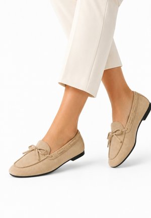 Mocasines beige de ante con detalle de lazo, usados con pantalones blancos recortados, mostrados con las piernas cruzadas sobre un fondo blanco.