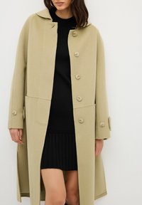 Cappotto di lana beige con un design strutturato, caratterizzato da cinque grandi bottoni, tasche laterali e un colletto arrotondato, indossato sopra un vestito a maglia nero.