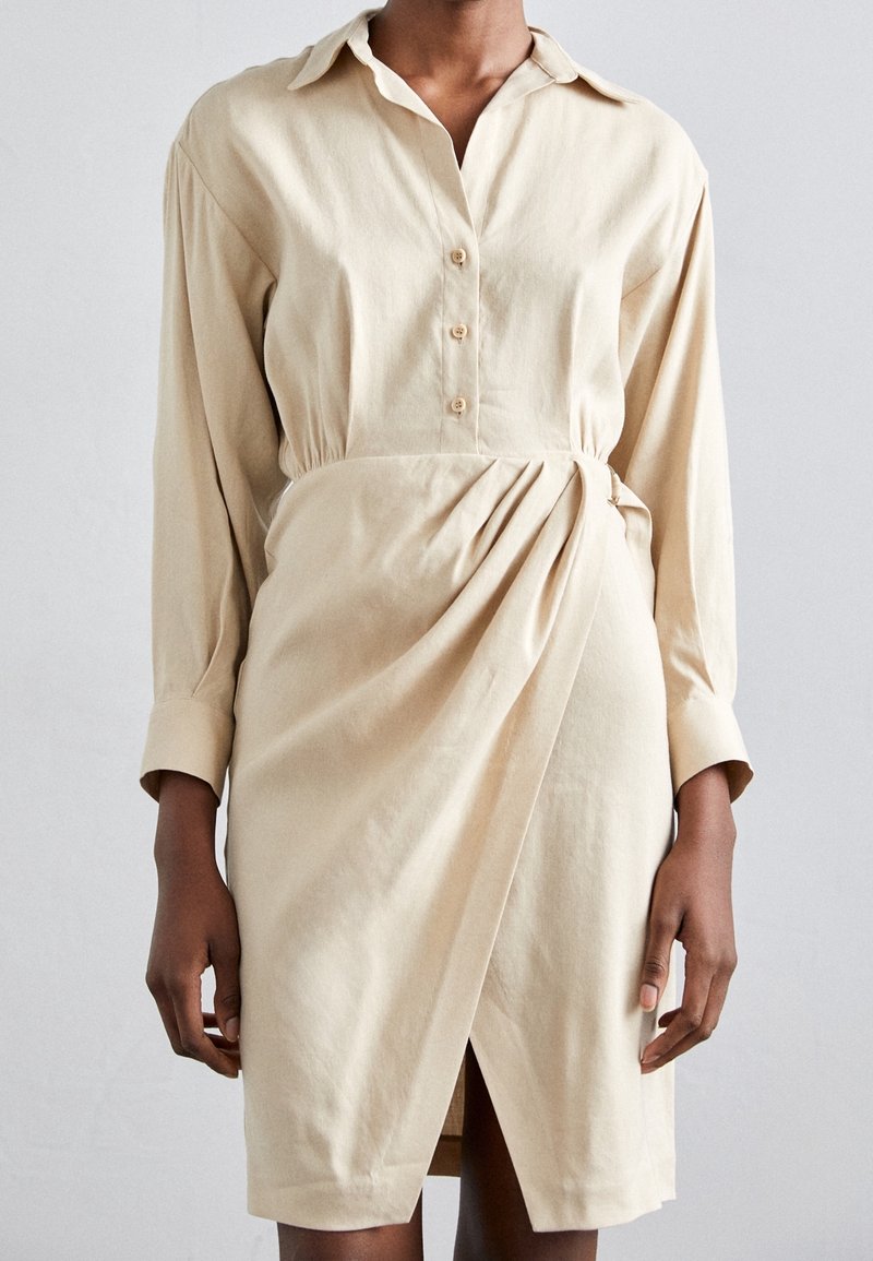 Personne portant une robe beige à manches longues boutonnée avec un devant de jupe drapé de style portefeuille et des larges poignets sur un fond uni.
