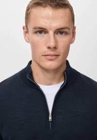 Marineblauer Reißverschluss-Pullover mit hohem Kragen, glatter Textur und silbernem Reißverschluss. Über einem weißen T-Shirt getragen. Nahaufnahme des Gesichts des Models.