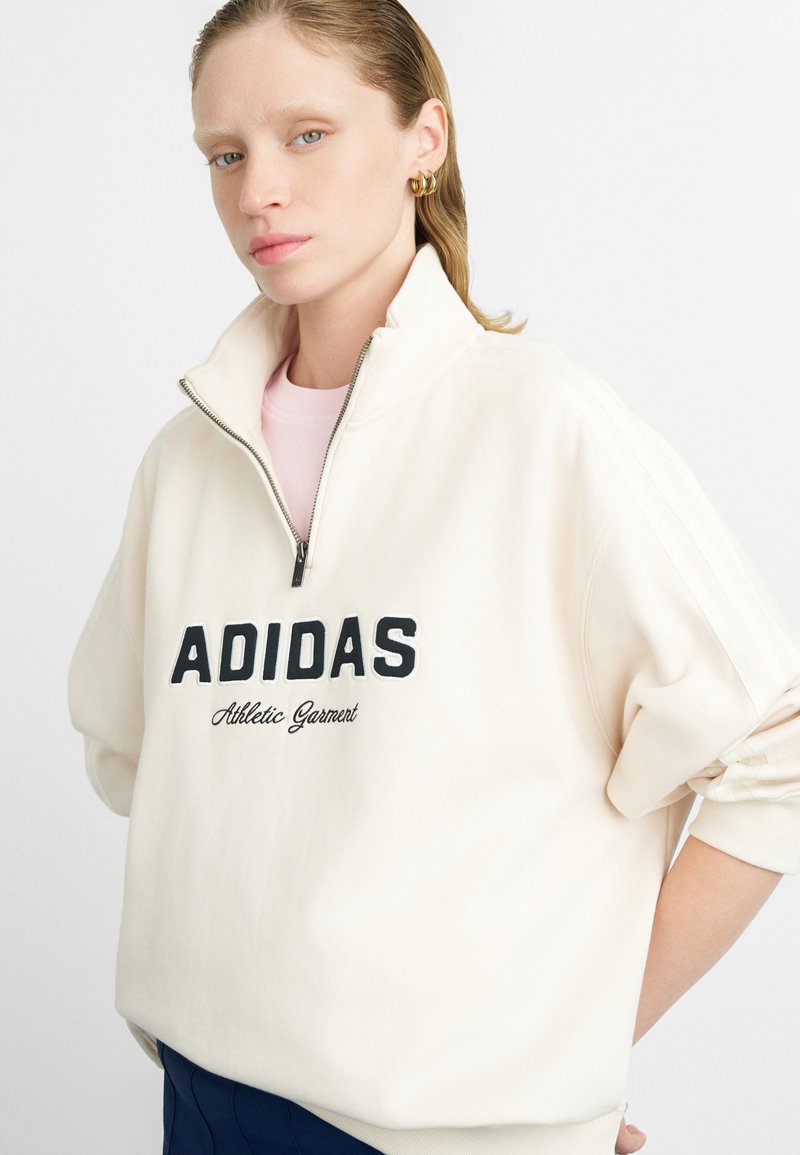 Felpa zip-up crema con collo alto, caratterizzata da un grande logo nero "ADIDAS" e dalla scritta "Athletic Garment". Texture morbida e vestibilità rilassata.