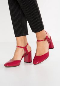 Chaussures à talons hauts rouges avec un bout pointu, une sangle à la cheville et un talon épais. Fini en cuir lisse avec des détails de texture subtils.