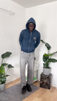 Jeans de perna larga cinzentos desbotados emparelhados com um hoodie azul-marinho com fecho e bolso frontal, equipado com cordões. Sapatos pretos completam o visual.
