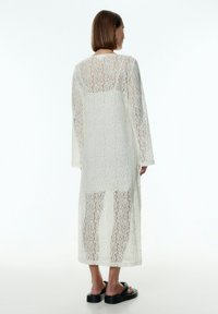 Langes, durchscheinendes weißes Spitzenkleid mit langen Ärmeln und lockerer Passform. Das kunstvolle Blumenmuster bedeckt das gesamte Kleidungsstück und zeigt eine glatte Innenschicht.