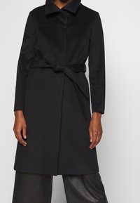 Černý kabát vyrobený z hladkého materiálu, s límečkem, předním zapínáním a uvázaným páskem v oblasti pasu. Dlouhé rukávy, rovný střih.