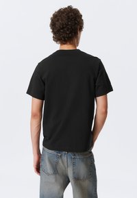 Black cotton t-shirt with short sleeves, crew neck, and a straight hem. Worn with light blue denim jeans featuring back pockets.