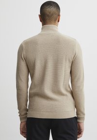 Beige pulóver magas nyakkal, texturált kötött mintázattal, bordázott mandzsettákkal és szegéllyel. Lazább szabásra tervezve.
