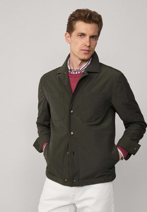 Dunkelgrüne Jacke aus glattem Stoff, mit zwei Fronttaschen, braunen Knöpfen und einem Kragen. Über einem roten Pullover und einem gestreiften Hemd getragen.