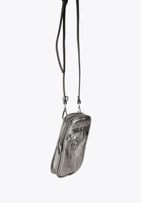 Malá metalická strieborná crossbody taška s embosovaným vzorom, predným zipsovým vreckom a nastaviteľným tenkým popruhom, visieť na jednoduchom pozadí.