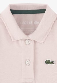 Ανοιχτό ροζ πόλο πουκάμισο με κοντό γιακά, υφή με ραβδώσεις, δύο λευκά κουμπιά και πράσινο κεντητό λογότυπο κροκοδείλου στο στήθος.