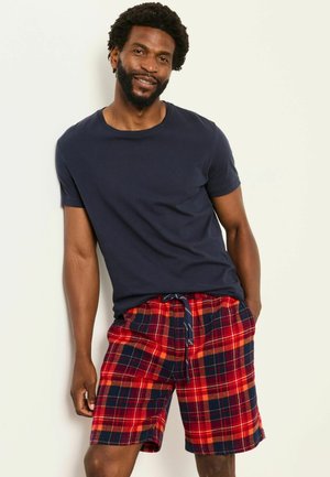 Mann mit kurzen lockigen Haaren und Bart, trägt ein dunkelblaues T-Shirt und rote karierte Shorts, steht mit den Händen in den Taschen vor einem hellen Hintergrund.