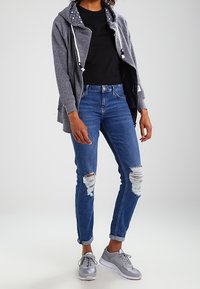 Felpa grigia con cappuccio e lacci sopra una t-shirt nera, abbinata a jeans blu strappati e sneaker grigie.