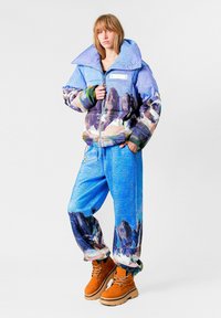 Chaqueta acolchada azul con un gran cuello y estampado de montaña; combinada con pantalones azules con patrones similares y botas color beige.