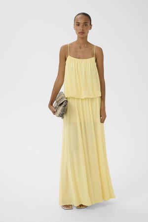 Femme debout portant une robe maxi fluide jaune pâle à fines bretelles, tenant une pochette en cuir de serpent avec des sandales.