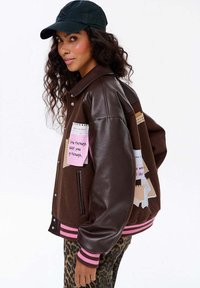 Kobieta w brązowej skórzanej kurtce varsity z różowymi łatami, w spodniach w panterkę i czapce bejsbolowej w kolorze czarnym, spogląda przez ramię.