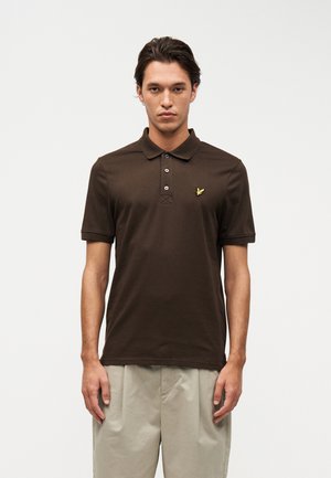 Jeune homme portant un polo à manches courtes marron avec un logo d'oiseau jaune sur la poitrine et un pantalon beige, debout devant un fond blanc.