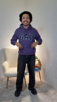 Nasmejan moški, ki nosi vijoličasto kapucarico GAP in črne hlače, stoji v prostoru z belo teksturirano stolom in majhno okroglo mizo, na kateri so knjige in vaza.