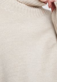 Maglione beige a collo alto in maglia, con un bordo a coste sul collo e sui polsini, che mette in mostra una texture morbida e una costruzione senza cuciture.