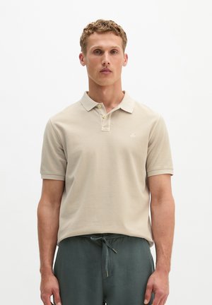 Junger Mann mit kurzem lockigem Haar, der ein beiges Poloshirt und graue Hose mit Kordelzug trägt, steht vor einem schlichten weißen Hintergrund.
