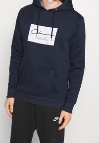 Felpa blu navy con una patch grafico bianco che presenta testo corsivo e "MODERN ESSENTIALS". Include una tasca frontale e cappuccio con cordino.