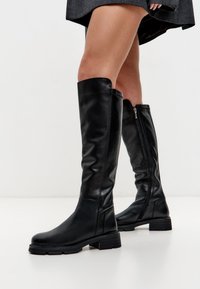 Botas de cuero negro hasta la rodilla con una textura suave, suela plana y una cremallera lateral para facilitar su uso. Detalles de costura sutil a lo largo de los bordes.