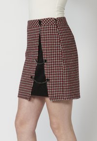 Jupe pied-de-poule rouge et noir avec une fente sur le côté, détail en chaîne et contraste de texture de tissu. Présente un design ajusté et des accents de boutons.