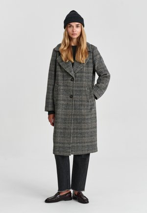 Femme portant un long manteau à carreaux gris, un bonnet noir, un jean court noir et des mocassins marron foncé, debout avec une main dans la poche.