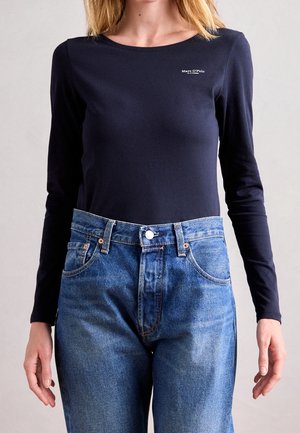 Femme portant un t-shirt bleu marine à manches longues avec le logo "Marc O'Polo" et un jean bleu taille haute, debout avec les bras détendus le long du corps.