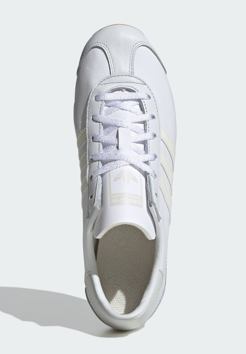 Zapatilla de piel blanca con punta redondeada, plantilla texturizada, tres franjas gris claro y cordones gris plata en la parte superior.