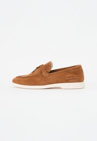 Mocasines de ante marrón con suela de goma blanca, costuras decorativas y un acento metálico plateado en la parte superior. Presenta una marca en el lateral.