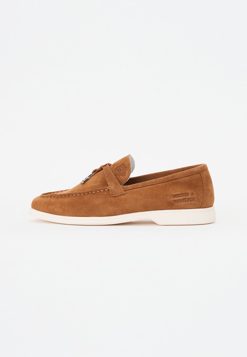 Mocasines de ante marrón con suela de goma blanca, costuras decorativas y un acento metálico plateado en la parte superior. Presenta una marca en el lateral.