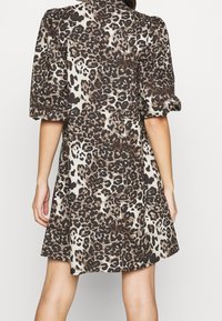 Kvinna som bär en kort klänning med leopardmönster och puffiga korta ärmar, sedd bakifrån mot en enfärgad bakgrund.