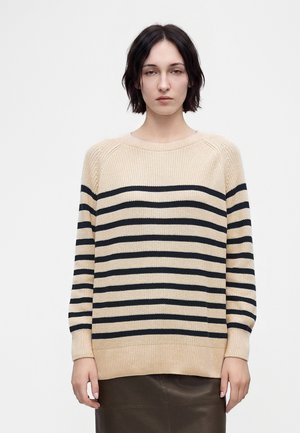 Vrouw met kort donker haar die een beige trui draagt met horizontale zwarte strepen en een donkere rok, staand tegen een effen lichte achtergrond.