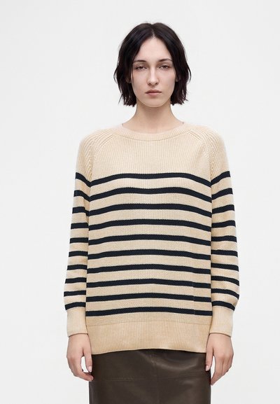 Femme aux cheveux courts et foncés portant un pull beige avec des rayures noires horizontales et une jupe foncée, debout devant un fond clair uni.