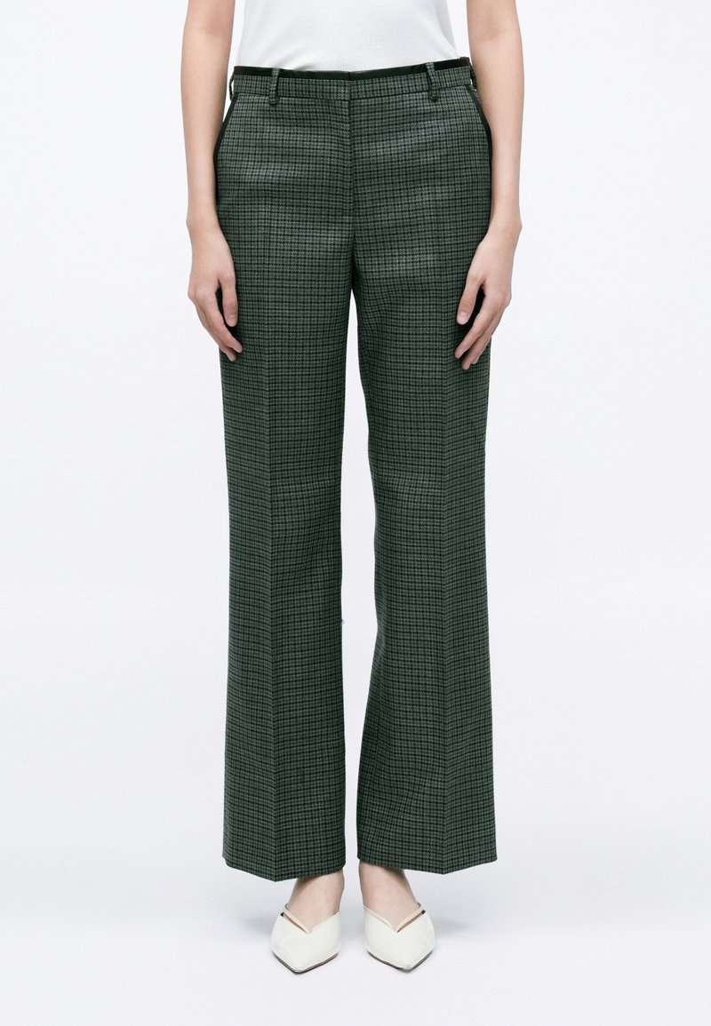 Femme portant un pantalon droit taille haute vert foncé à carreaux avec des poches et des chaussures blanches à bout pointu sur fond blanc.