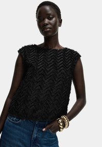 Top sin mangas negro texturizado con un patrón de chevron, hecho de un material borroso. Combinado con una pulsera de bangles dorada y pantalones de mezclilla.