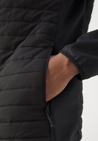 Sort jakke med en quiltet sektion med vandrette linjer og en sidelomme. Stoffet ser glat ud med en mat finish.
