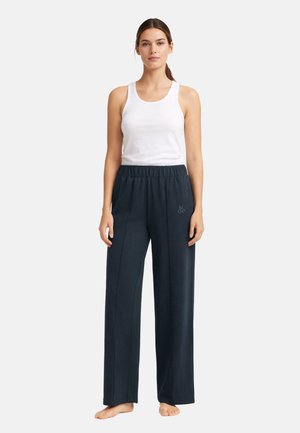 Frau steht barfuß und trägt ein weißes ärmelloses Oberteil sowie lockere, dunkelblaue Weite-Hosen mit elastischem Bund und kleinem Logo.