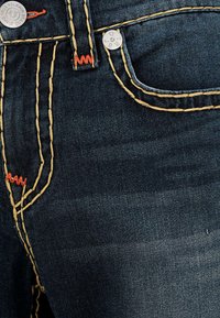 Jeans de mezclilla oscura con costuras amarillas y rojas, un botón plateado y un bolsillo delantero. La tela tiene un acabado liso y ligeramente texturado.