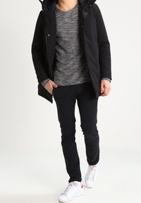 Man som bär svart kappa, grå stickad tröja, svarta byxor och vita sneakers, går med händerna i fickorna.