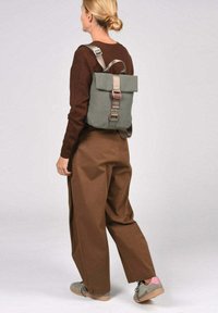 Sac à dos en tissu vert avec un haut roulé et des bretelles réglables, doté d'une boucle grise et d'un accent rouge. Porte sur un pantalon marron à jambes larges.