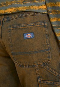 Rudos džinsinio audinio kelnes su išskirtiniu kišenės dizainu. Audinys turi tekstūrinį paviršių, o gale matomas Dickies logotipo pleistras.